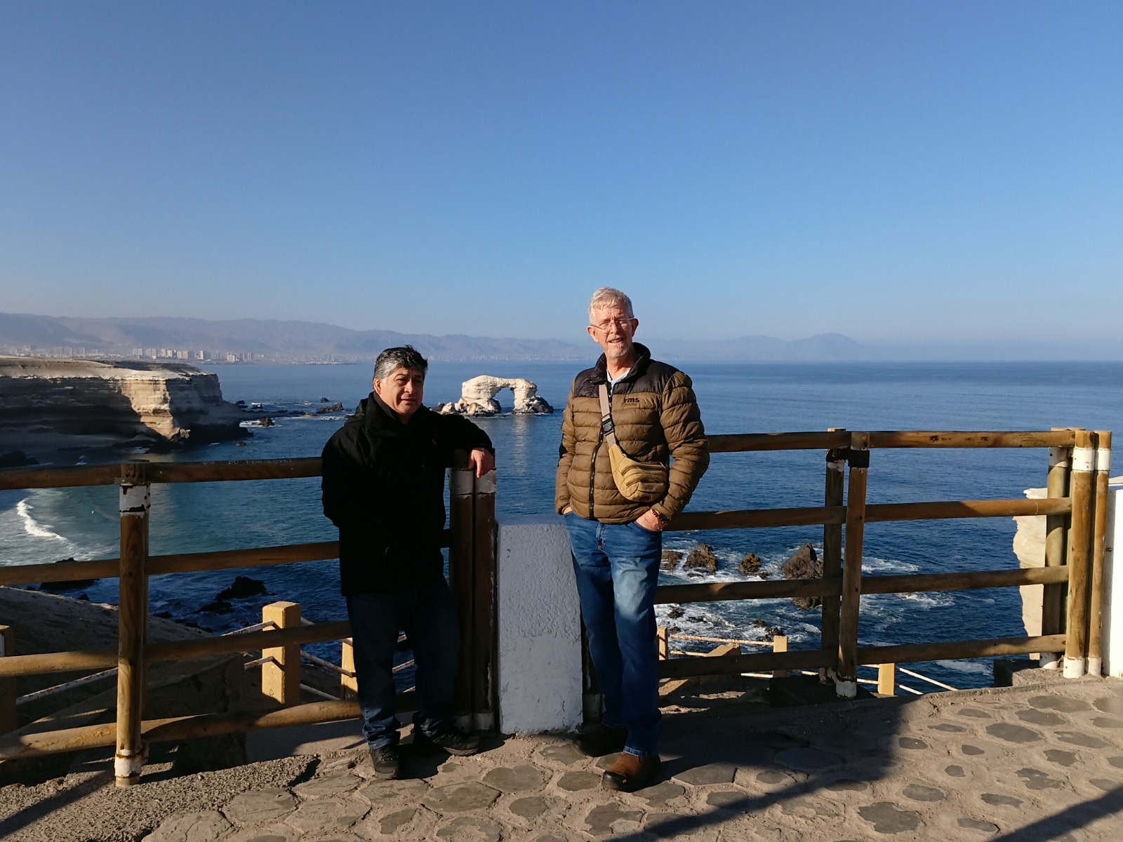 Two people at a coastal viewpoint overlooking the ocean and a natural rock arch in Chile